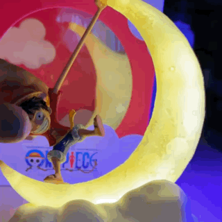 A close-up animated view of the One Piece Luffy Gear 5 figure on an illuminated crescent moon lamp, with the background cycling between a pink/blue cloud effect and a dark night setting to show the dual display modes