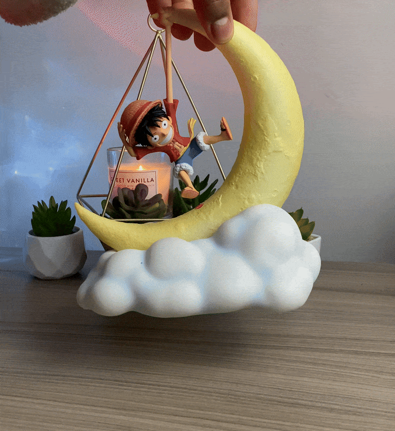 A hand demonstrates the dynamic setup of the One Piece Luffy Gear 5 figure sitting on a glowing crescent moon lamp, positioned next to a decorative terrarium candle