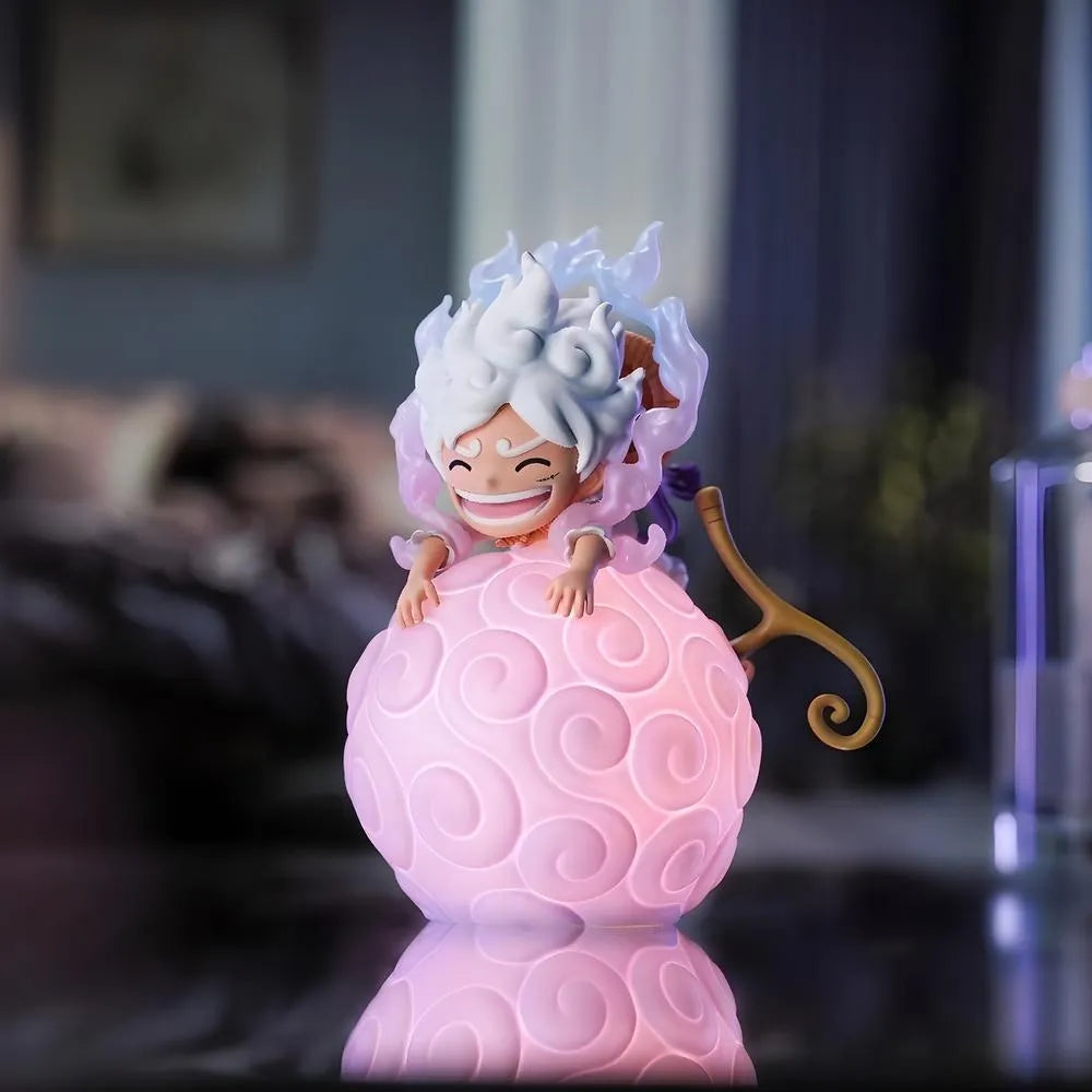 A front-facing view of a Luffy Gear 5 Nika figure, with his Straw Hat on his back, sitting happily on a large, glowing pink One Piece devil fruit night light in a room setting